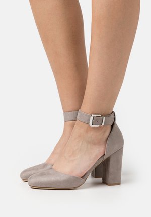 Jambes portant des chaussures à talons hauts en daim taupe avec des brides à la cheville et des boucles argentées, posées sur une surface blanche.