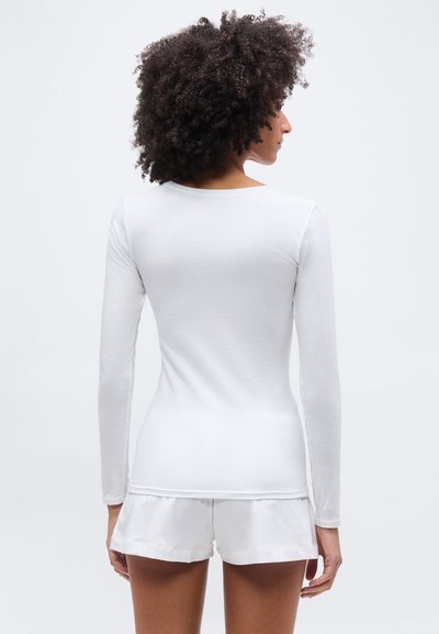 Άτομο με κατσαρά μαλλιά που φαίνεται από πίσω, φοράει εφαρμοστή λευκή μακρυμάνικη μπλούζα και λευκά σορτς, μπροστά σε απλό φόντο.