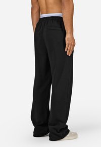 Calças de treino pretas com um corte descontraído, cós elástico e um detalhe de bolso nas costas. Tecido macio e texturizado; usadas com sapatos de cor clara.