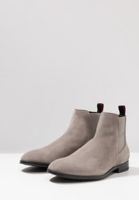 Une paire de bottes Chelsea en cuir suédé gris avec des semelles noires et des tirants accents rouges, présentée sur une surface blanche avec un arrière-plan gris clair.