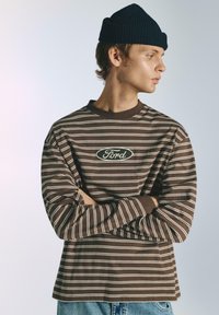 Barna hosszú ujjú csíkos ing krémszínű vonalakkal, kerek nyakkivágással és központi ovális "Ford" logóval. Sötét sapkával viselve.