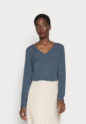 Blusa a maniche lunghe con scollo a V profondo in blu ardesia, realizzata in tessuto leggero, abbinata a una gonna midi plissettata color avorio.