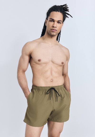 Muscular man with long dreadlocks wearing olive green swim shorts, standing with hands in pockets against a plain light background.