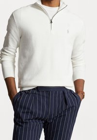 Vit stickad tröja med kvartdels dragkedja och ribbade ärmar. Har en diskret logotyp på bröstet. Kombinerad med marinblå randiga byxor.