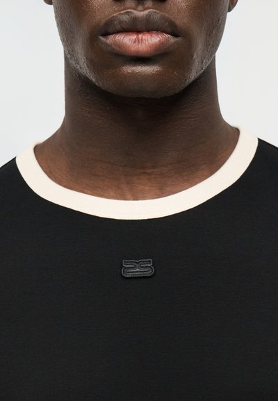 Κοντινό πλάνο ενός ατόμου που φοράει μαύρο πουκάμισο με λευκό γιακά και ένα μικρό μαύρο κεντητό λογότυπο στο κέντρο κάτω από το λαιμό.