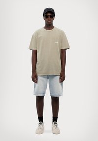 T-shirt oversized verde-claro com um pequeno logótipo branco, combinado com calções de ganga azul-claro, meias pretas e ténis bege. Boné preto e óculos de sol.