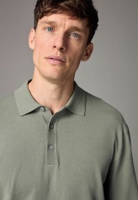 Polo de culoare verde deschis, realizat dintr-un material tricotat, având un guler, două butoane gri și detalii dublate pe guler și plăcuța butonului.
