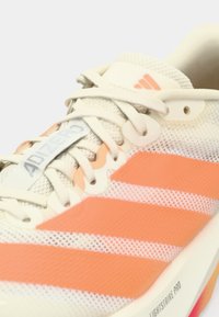 Lehká běžecká obuv s krémovým síťovaným svrškem, oranžovými pruhy, značkovým jazykem a texturovanou podešví s živými akcenty.