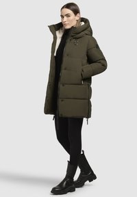Geacă puffer de culoare verde măslinie, cu guler înalt, căptușită cu blană sintetică, cu închidere cu fermoar și nasturi, și buzunare laterale. Botine negre completează ținuta.