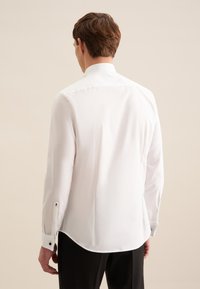 Man van achteren gezien, draagt een wit overhemd met lange mouwen en een zwarte broek tegen een effen beige achtergrond.