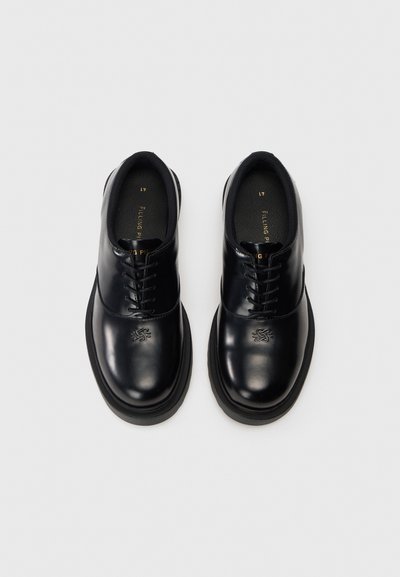 Schwarze lackierte Schnürschuhe mit runder Kappe, Gummisohle und eingeprägtem Logo auf dem Blatt. Glatte Textur und klares Design.