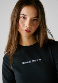 T-shirt noire à manches longues avec col rond, arborant un texte brodé blanc "RED BULL RACING" sur le devant. Texture lisse, design ajusté.