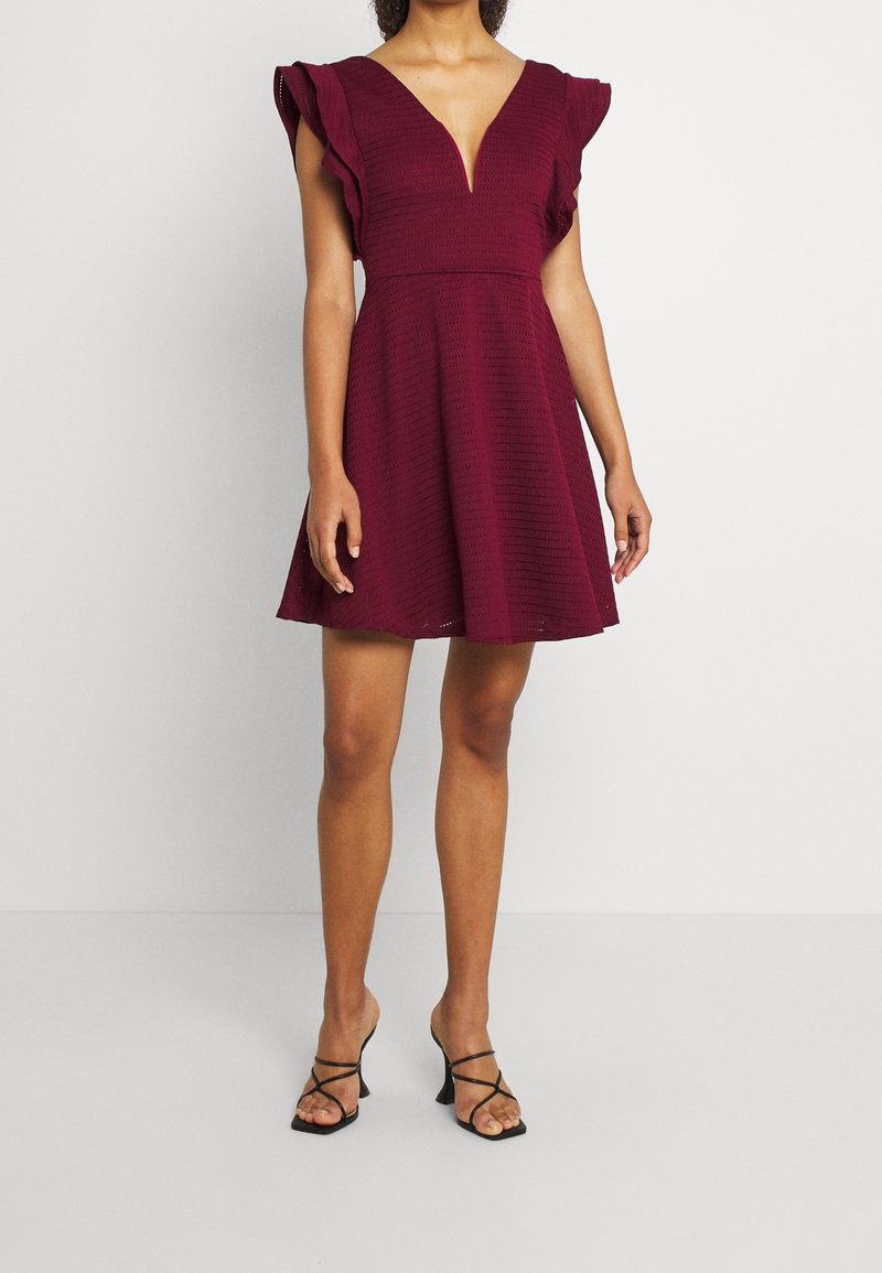 Femme portant une robe bordeaux longueur genou avec des manches volantées et un profond décolleté en V, associée à des sandales noires à talons avec brides.