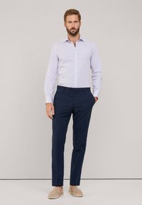 Camicia azzurra a righe bianche con bottoni, pantaloni blu scuri con frontale piatto e scarpe beige slip-on, modello in piedi contro uno sfondo neutro.