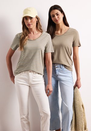 Dwie kobiety stoją, jedna w beżowej, w paski koszulce, białych spodniach i żółtej czapce; druga w jednolitym beżowym t-shircie, jasnoniebieskich dżinsach, trzymająca dzianinowy sweter.