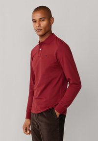 Polo rouge à manches longues avec col classique, patte de boutonnage à deux boutons et détail brodé sur la poitrine, associé à un pantalon foncé.