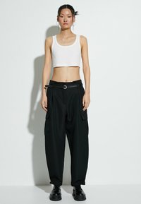 Witte geribde crop top, zwarte cargobroek met zakken, zwarte riem met een ronde gesp en dikke zwarte schoenen, gepresenteerd tegen een effen achtergrond.