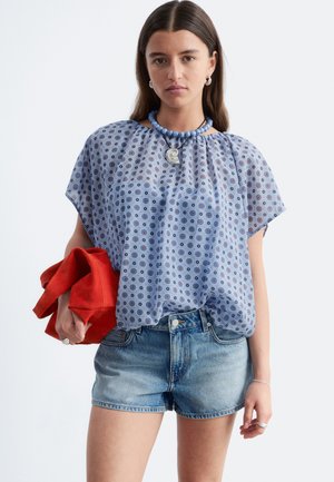 Femme portant un chemisier ample bleu à motifs, un short en jean, un collier de perles bleu, un pendentif en argent, tenant une veste rouge pliée dans sa main gauche.