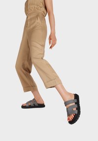 Sandali in tessuto grigio con suole nere, caratterizzati da due fasce larghe sulla parte superiore del piede e dettagli texture. Indossati con pantaloni cropped beige.