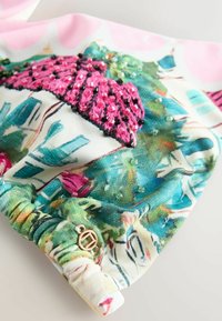 Közelkép színes anyagról zöld, kék és rózsaszín mintákkal, rózsaszín és fekete gyöngyös hímzéssel, valamint egy kis arany, kör alakú medállal.