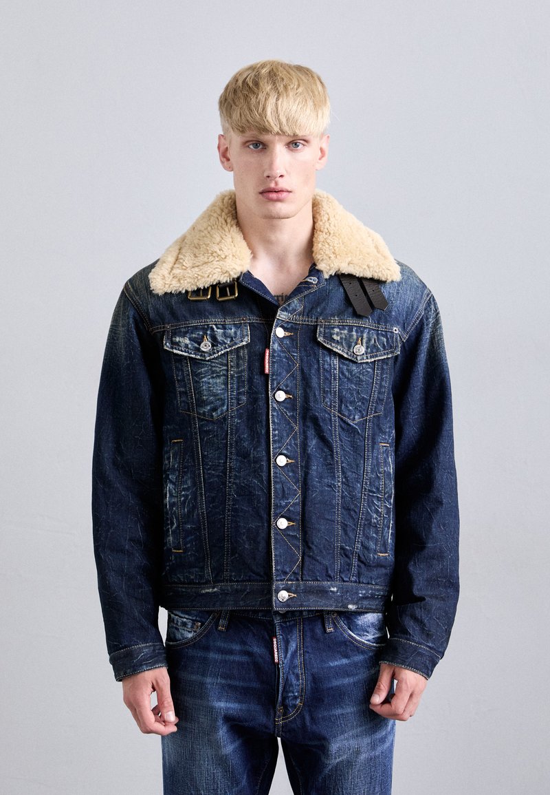 Denim jacket with a tan sherpa collar, front pockets, and metal buttons. The fabric shows a distressed texture and a dark blue color.