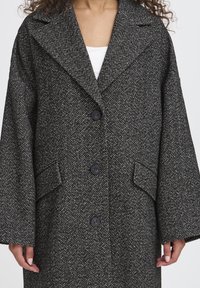 Kvinde iført en mørkegrå hørhammert coat med brede revers, tre sorte knapper og klaffelommer over en hvid top.