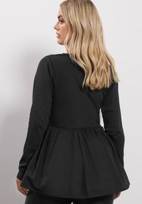 Pitkähihainen musta paita, jossa on istuva vartalo ja liehuva peplum-helma. Ominaisuuksina sileä pinta ja uritettu yksityiskohta hihoissa.