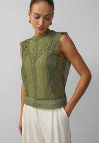 Olivgrünes Spitzenoberteil mit gewellten Kanten, aufwendigen Blumenmustern und einer strukturierten Oberfläche. Mit tailliertem Oberteil und ärmellosem Design.