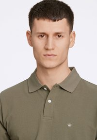 Olijfgroene polo shirt van gestructureerde stof; beschikt over een klassieke kraag, drie knopen en een klein geborduurd logo op de linkerborst.