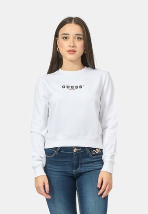 Felpa bianca corta con il logo "GUESS?" in testo nero e rosso. Tessuto morbido con maniche lunghe e polsini a coste, abbinata a jeans blu.