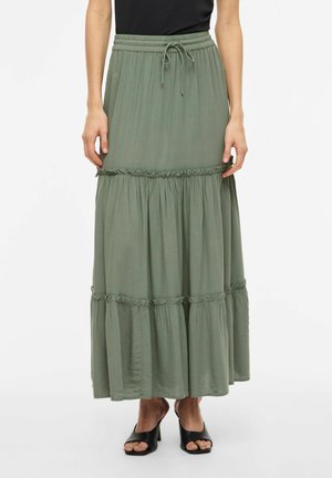 Žena v dlhom olivovo zelenom sukni s volánmi a zaväzovaním v páse, s čiernymi otvorenými sandálmi na podpätku, stojaca na bielom pozadí.