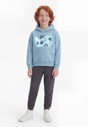 Enfant aux cheveux roux debout, portant un sweat à capuche bleu clair avec un imprimé de panier de basket, un pantalon gris foncé et des baskets montantes blanches.