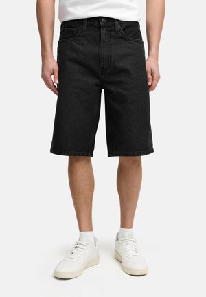 Man draagt zwarte denim knielange shorts, witte sokken en witte sneakers, staand tegen een effen witte achtergrond.