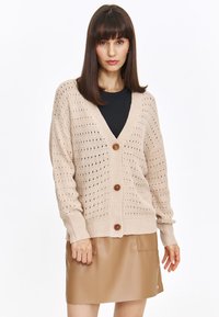 Sweter kardigan w miękkim beżowym kolorze z otwartym splotem, z dekoltem w literę V i pięcioma dużymi brązowymi guzikami. Połączony z gładką brązową spódnicą.