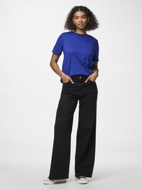 Kvinna med lockigt hår som bär en blå t-shirt med kort ärm, svarta jeans med vida ben och vita sneakers, stående med händerna i fickorna.