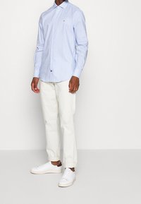 Hellblau kariertes Hemd mit langenÄrmeln, kombiniert mit weißen Stoffhosen und weißen Turnschuhen. Das Hemd hat ein kleines Logo auf der Brust.