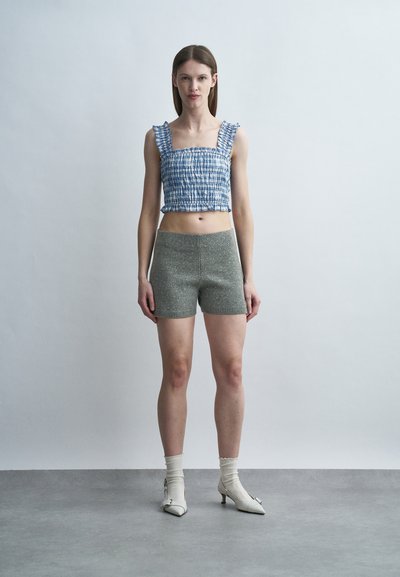 Νεαρή γυναίκα όρθια, φοράει μπλε τοπ με βολάν, γκρι σορτς με στίγματα, λευκές κάλτσες και λευκά παπούτσια με τακούνι kitten, σε απλό φόντο.