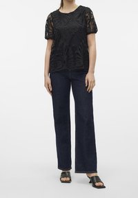 Top en dentelle noire avec des motifs floraux complexes, manches courtes, associé à un pantalon en denim foncé taille haute à jambes évasées et des sandales noires à enfiler.