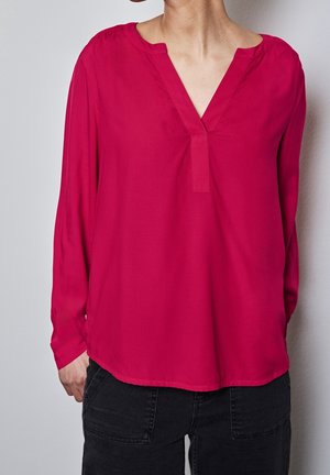 Vrouw draagt een lange mouw, dieproze V-hals blouse gecombineerd met een zwarte broek, staand tegen een effen lichtgrijze achtergrond.