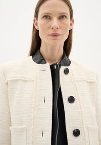 Giacca bianca testurizzata con colletto in pelle nera, caratterizzata da due grandi bottoni e una zip. Dettagli significativi includono tasche e bordi sfrangiati.