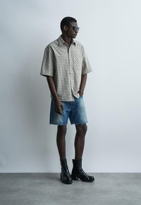 Homme portant des lunettes de soleil, une chemise à manches courtes à carreaux, un short en jean et des bottines noires à talons, debout contre un mur uni.