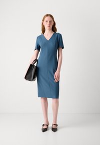 Blue short-sleeve dress with a V-neck, textured fabric, and seam detailing. Paired with a structured black handbag and pointed flats.