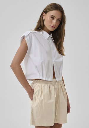 Femme portant un crop top blanc sans manches à col et un short beige taille haute avec cordon, debout avec une main dans la poche devant un fond gris.