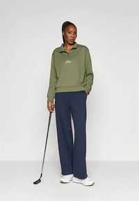 Olivengrøn sweatshirt met een halve rits en logo, gecombineerd met marineblauwe wijde broek. Model staat, houdt een golfclub vast en draagt witte sneakers.