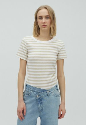 Frau mit schulterlangen blonden Haaren, die ein weißes und beige gestreiftes T-Shirt und asymmetrische blaue Jeans vor einem einfachen Hintergrund trägt.