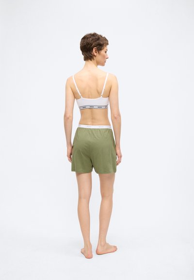 Femme debout pieds nus, de dos, portant un brassière de sport blanche marquée et un short large vert olive sur un fond blanc uni.
