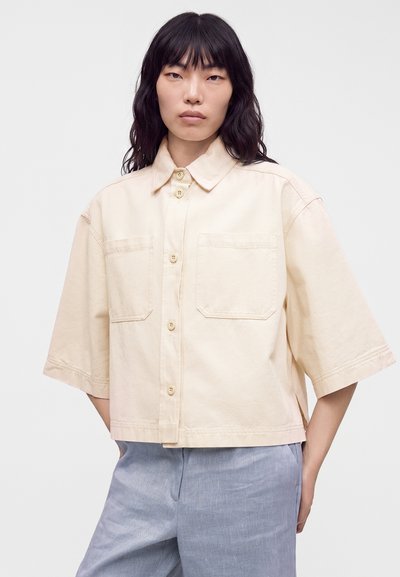 Femme aux cheveux noirs mi-longs portant une chemise beige ample boutonnée avec deux poches poitrine et un pantalon gris clair.
