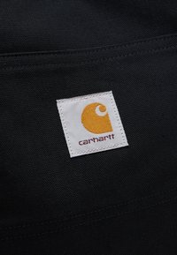 Kvadratna bela tkaninska etiketa z rumenim logotipom Carhartt in rjavim besedilom na teksturiranem črnem tkanem ozadju.