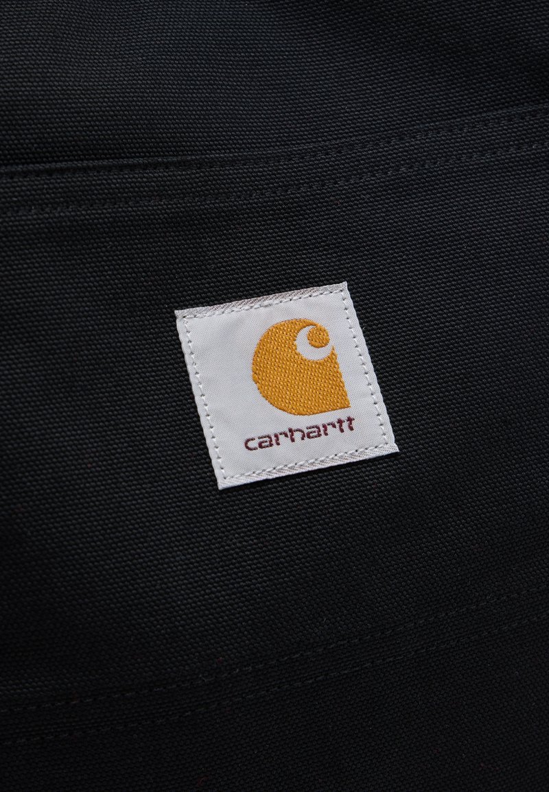 Square white fabric label with yellow Carhartt logo and brown text on textured black fabric background.