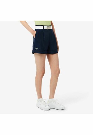 Persona che indossa pantaloncini blu navy con cintura bianca, scarpe da ginnastica bianche e top verde chiaro, in piedi davanti a uno sfondo bianco semplice.